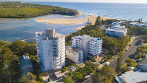 Vue aérienne d’un quartier résidentiel situé sur la côte, avec des propriétés en bord de mer, notamment des immeubles à plusieurs étages dotés de balcons, et une plage en arrière-plan.