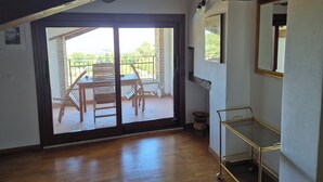 Una habitación con suelo de madera, una puerta corrediza de cristal que da a un balcón, un estante metálico y un espejo en la pared.