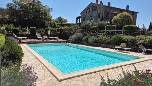 Una piscina rectangular con borde de piedra, rodeada por un patio con baldosas de piedra. Hay sillones de descanso y una sombrilla.