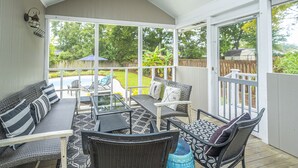 Eine überdachte Terrasse mit Korbmöbeln, einem Glastisch und Blick auf einen Garten mit Holzzaun und viel Grün.