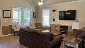 A living room with a brown sectional sofa, two armchairs, a wooden coffee table, a TV mounted on the wall, and a ceiling fan. - Escape the heat and cool off this summer in the Guadalupe! (New Braunfels)
