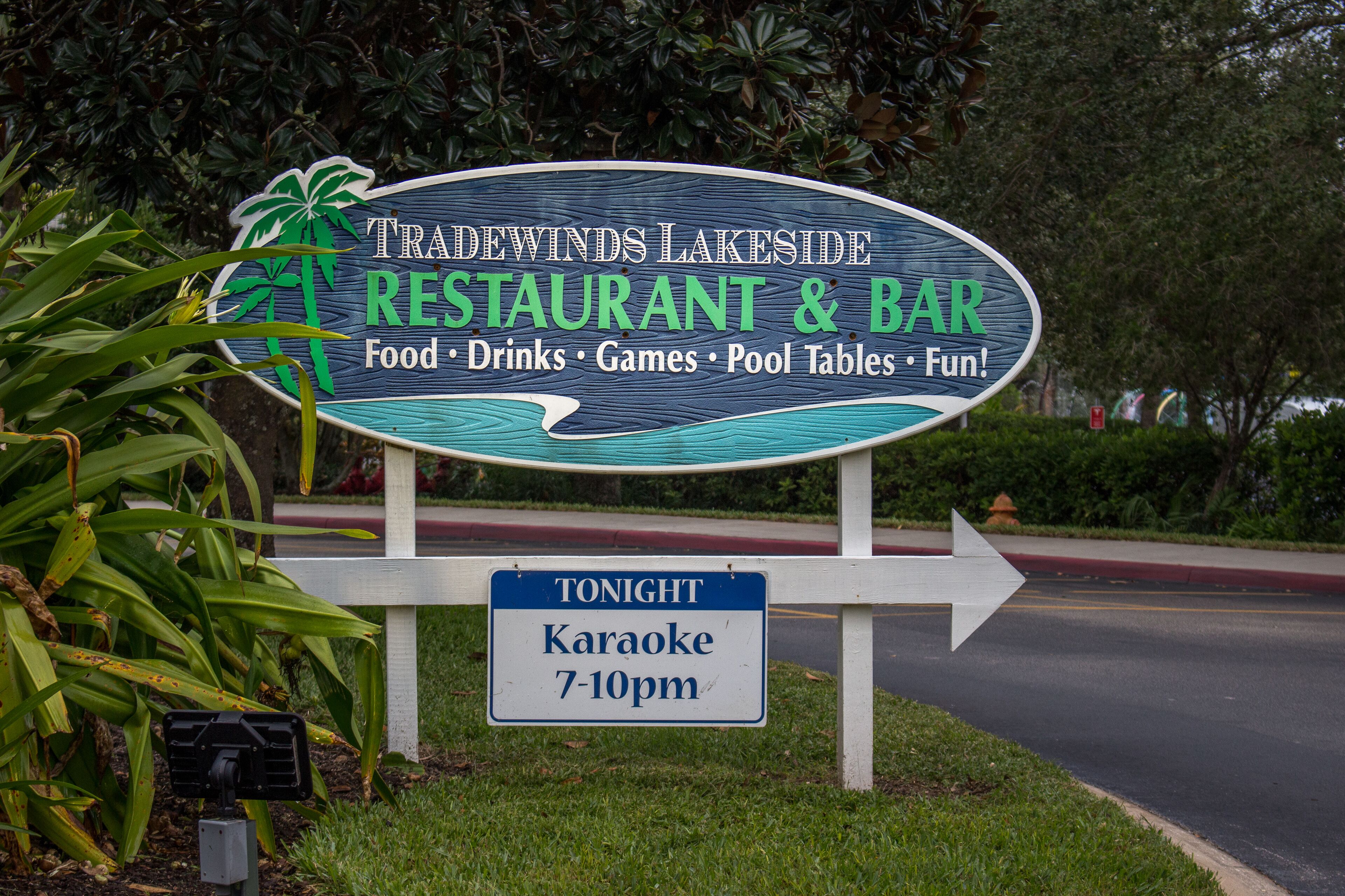 Une enseigne indique les services offerts par le restaurant-bar Tradewinds Lakeside, notamment la nourriture, les boissons, les jeux, les tables de billard et les activités de loisirs. Elle annonce également un événement karaoké ce soir, de 19 h à 22 h.