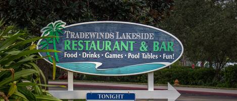Une enseigne indique les services offerts par le restaurant-bar Tradewinds Lakeside, notamment la nourriture, les boissons, les jeux, les tables de billard et les activités de loisirs. Elle annonce également un événement karaoké ce soir, de 19 h à 22 h.