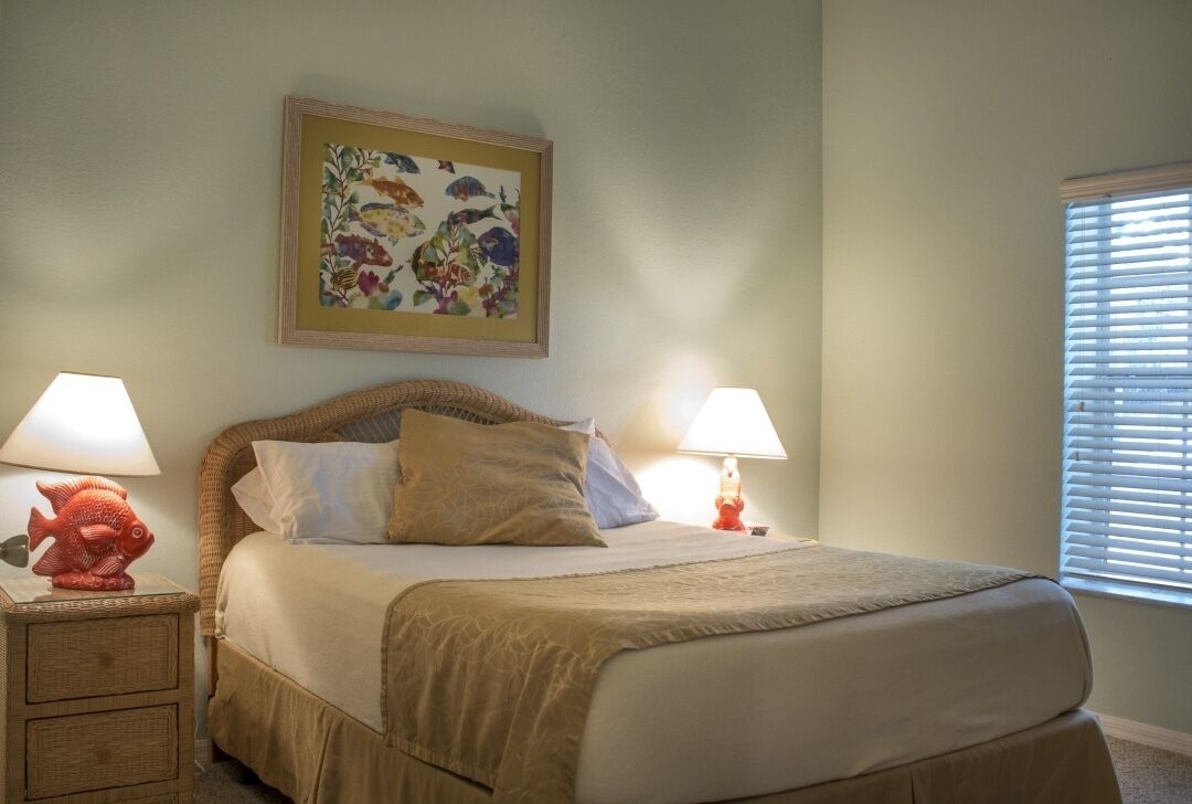 Una cama bien hecha con una colcha color beige y almohadones, flanqueada por dos mesitas de noche con lámparas. Encima de la cama, un cuadro enmarcado con motivos florales.