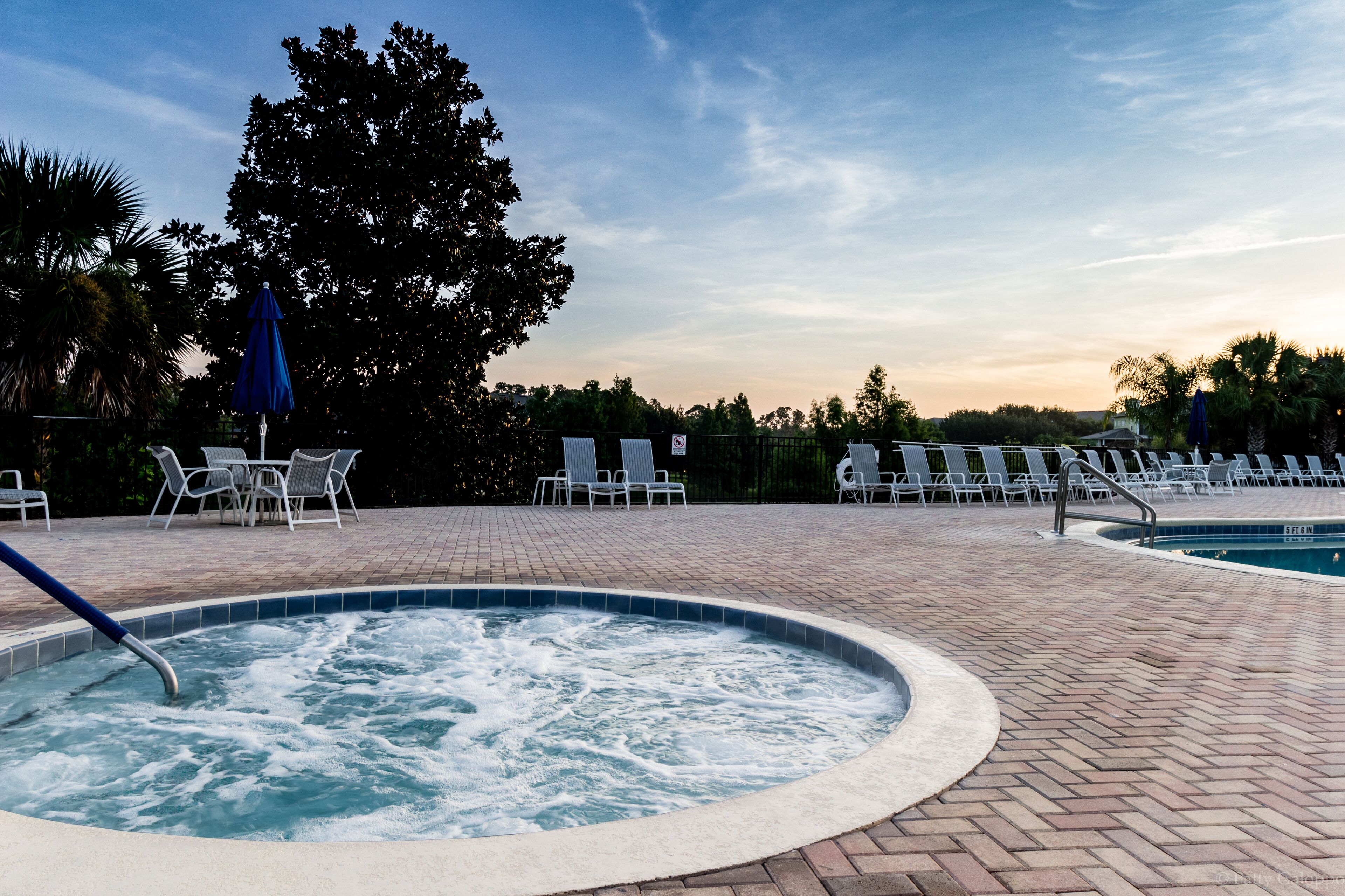 Un bain à remous entouré de chaises longues et une piscine, avec des arbres et un ciel dégagé en arrière-plan.