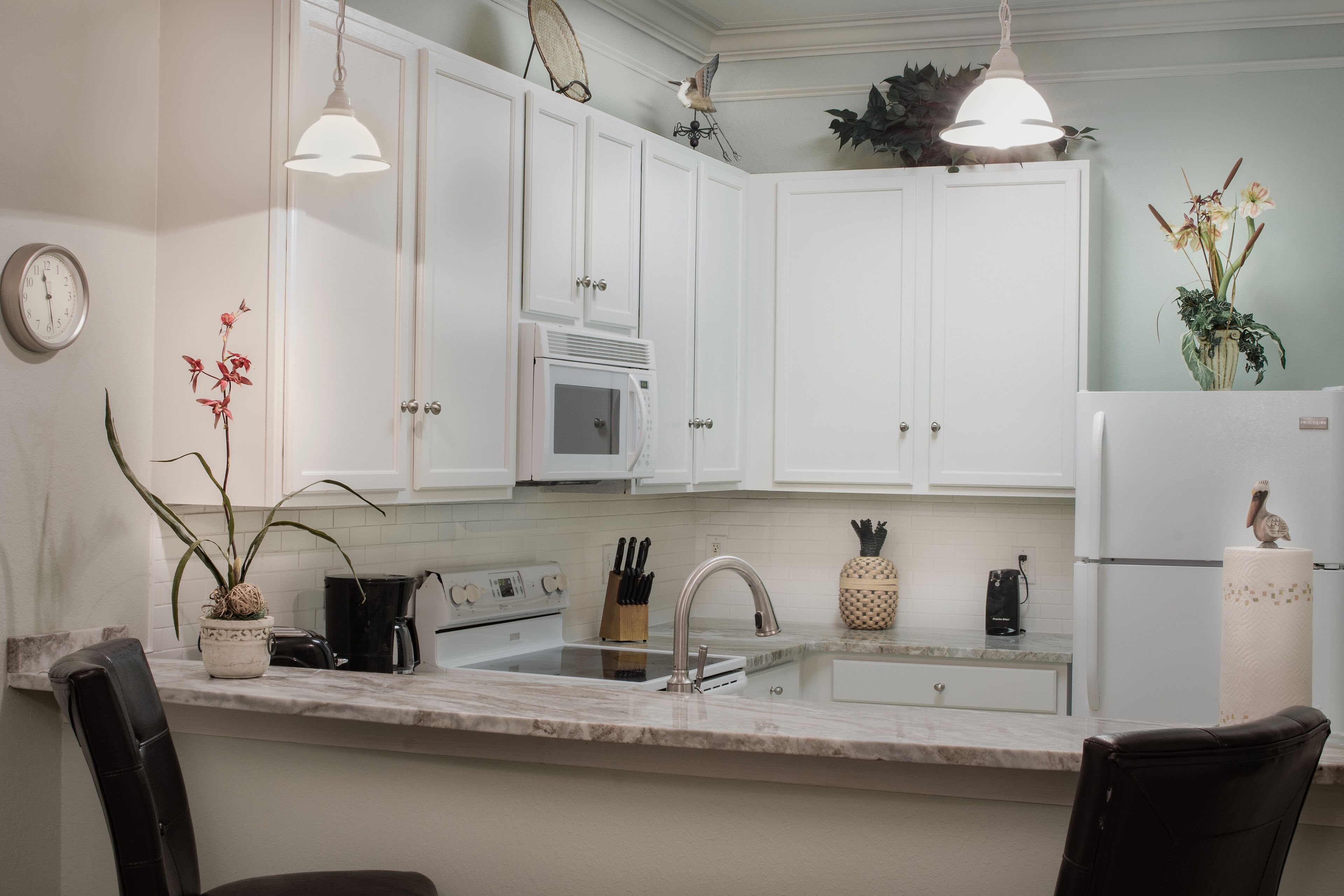 A modern kitchen with white cabinets, a marble countertop, and a built-in oven. There are two pendant lights, a clock, and a vase with flowers on the wall.