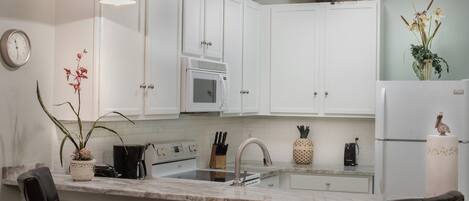 A modern kitchen with white cabinets, a marble countertop, and a built-in oven. There are two pendant lights, a clock, and a vase with flowers on the wall.
