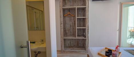 A hotel room with a wooden wardrobe, a small bathroom with a mirror, and a bedside table with a tray of toiletries.
