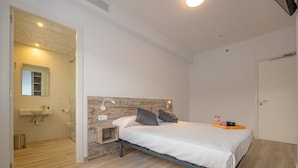 Een slaapkamer met een bed, nachtkastje en bijzettafeltje. Er is een badkamer met een wastafel en spiegel. De kamer heeft houten vloeren en witte wanden.