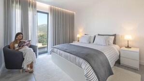 A modern bedroom with a large bed, two bedside tables with lamps, a chair, and a view of the outdoors through large windows. - Longra Family Villa & SPA (Mirandela)