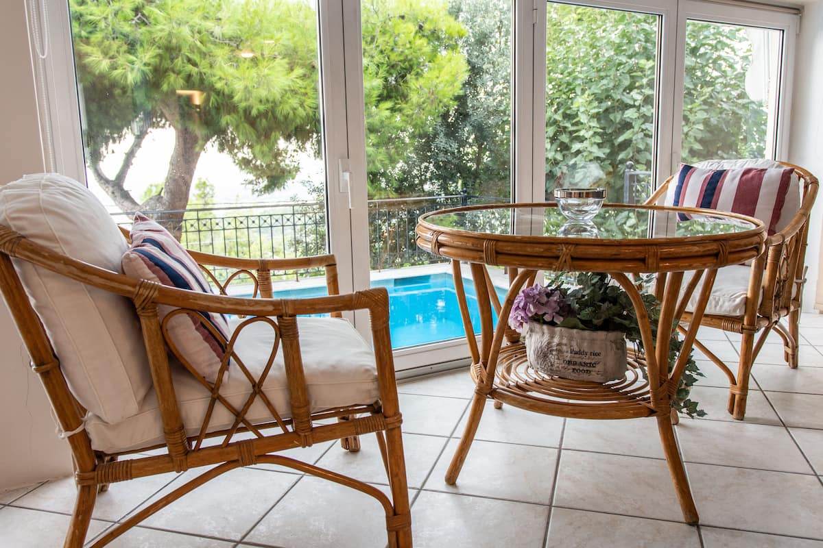 A wicker chair with a cushion and a striped pillow, a glass table with a vase of flowers, and a view of a swimming pool through large windows.