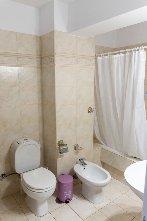 Une salle de bain avec des carreaux de couleur beige, des toilettes, un bidet, une poubelle et un rideau de douche.