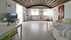 A spacious living room with a wooden ceiling, a television, a bookshelf, and a window with a view of greenery. - Apartment Anker am See (Wangen)