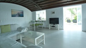 A spacious living area with a white sofa, a coffee table, and a television on a stand. There is a dining table with chairs and a kitchen area visible through an open door. - Apartment Anker am See (Wangen)
