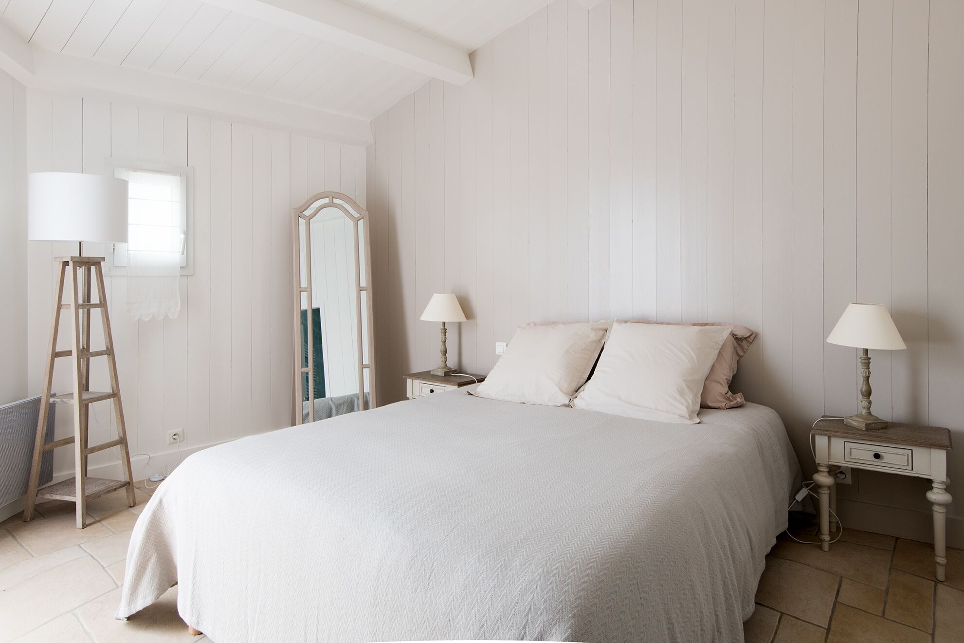A bedroom with a large bed, two bedside tables with lamps, a wooden ladder, and a mirror.