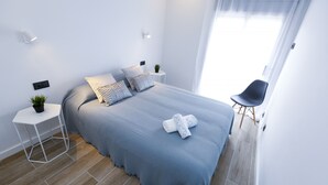 A modern bedroom with a neatly made bed, a small side table with a plant, and a chair by the window. - SOL DE ESPAÑA Apartments - Beachfront (Cambrils)