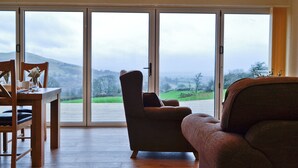 A living room with a wooden floor, a sofa, a dining table with chairs, and large glass doors leading to a view of a green landscape.