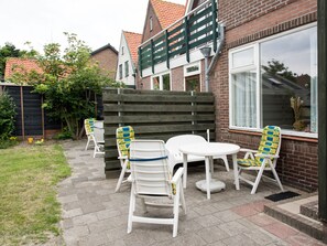 A patio with white plastic chairs and a table, a wooden fence, and a brick building with a window.