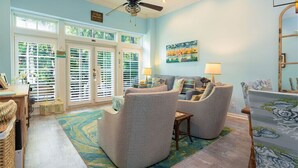 A spacious living room with a ceiling fan, large windows with blinds, a colorful rug, and a painting on the wall. - Hemingway Hideaway in the Truman Annex (Key West)