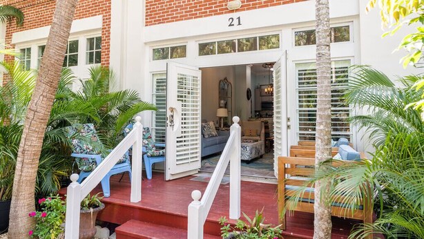 A brick building with a number 21 sign, a staircase leading to a porch with blue chairs, and a view into the interior with a sofa and lamp. - Hemingway Hideaway in the Truman Annex (Key West)