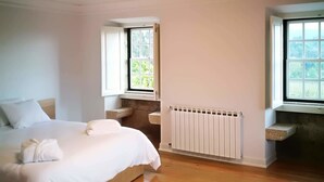 A modern bedroom with a wooden floor, a large bed with white linens, a built-in bench, a radiator, and a window with a view of greenery. - Solar das Bouças (Amares)