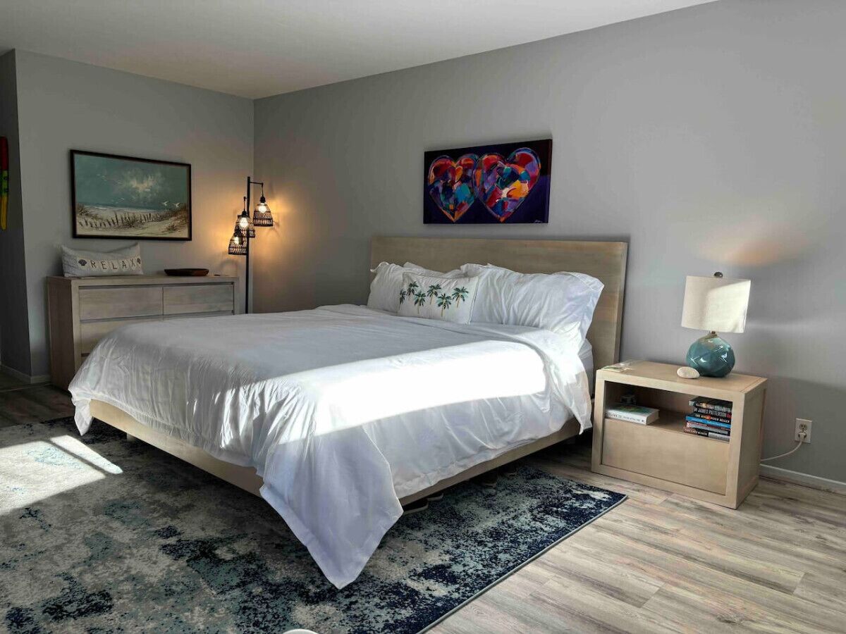 Une chambre à coucher moderne avec un grand lit, une table de chevet avec une lampe, une petite bibliothèque et des œuvres d’art accrochées aux murs.