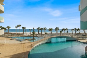 A pool area with a bridge leading to a beach, surrounded by palm trees and a clear sky.