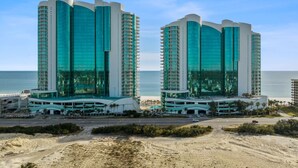 Et hotel med to spejllignende bygninger, udsigt til stranden og en parkeringsplads.