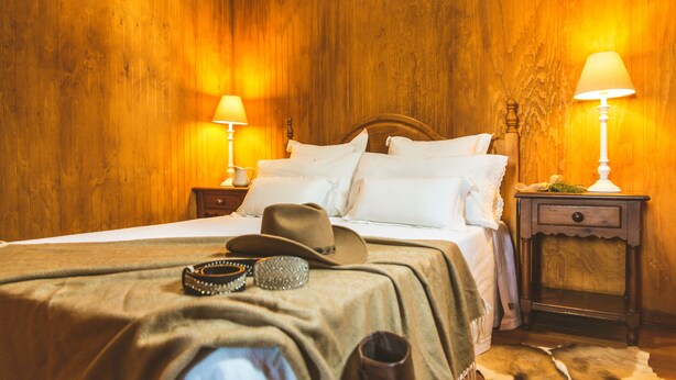 A wooden-paneled room with a bed, two nightstands, a lamp, a hat, and a belt on the bed. - Camping Fort Bravo (Tabernas)