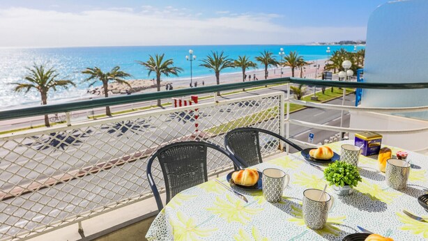 A balcony with a table set for dining, overlooking a beach with palm trees and the sea.