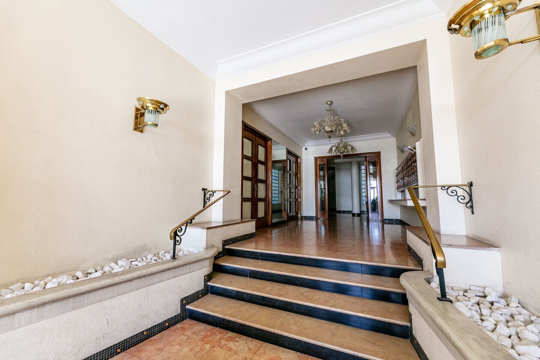 A grand staircase with marble steps and a handrail, leading to a hallway with a chandelier and a view of another room.