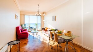 A bright living room with a red chair, a glass dining table, and a view of the sea through large windows.