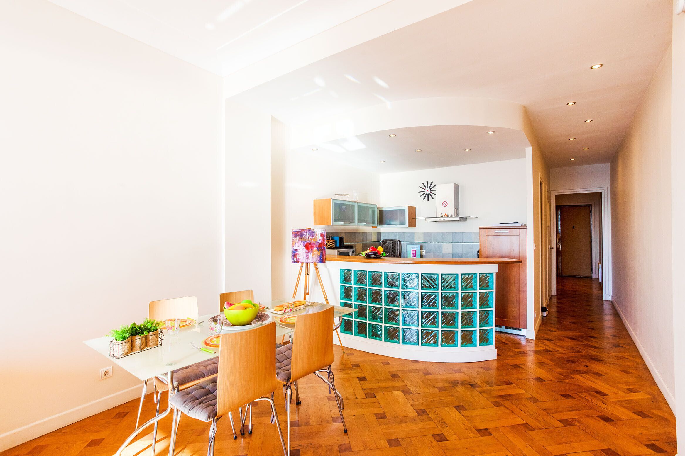 Une cuisine moderne dotée d’un îlot central aux lignes courbes, d’un plancher en bois et d’un coin repas avec une table et des chaises en verre.