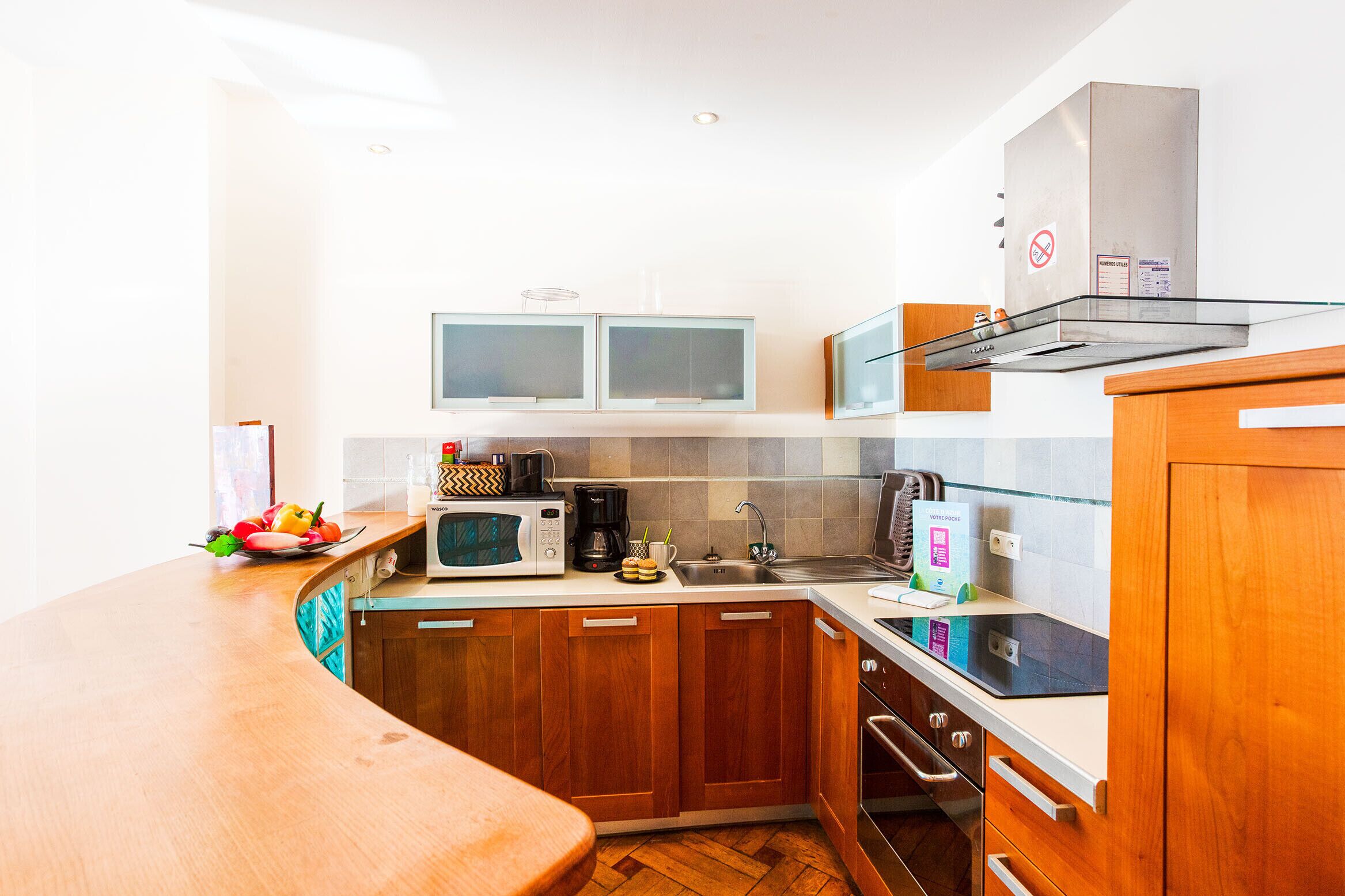 A compact kitchen with wooden cabinets, a microwave, a coffee maker, and a glass-top dining table.