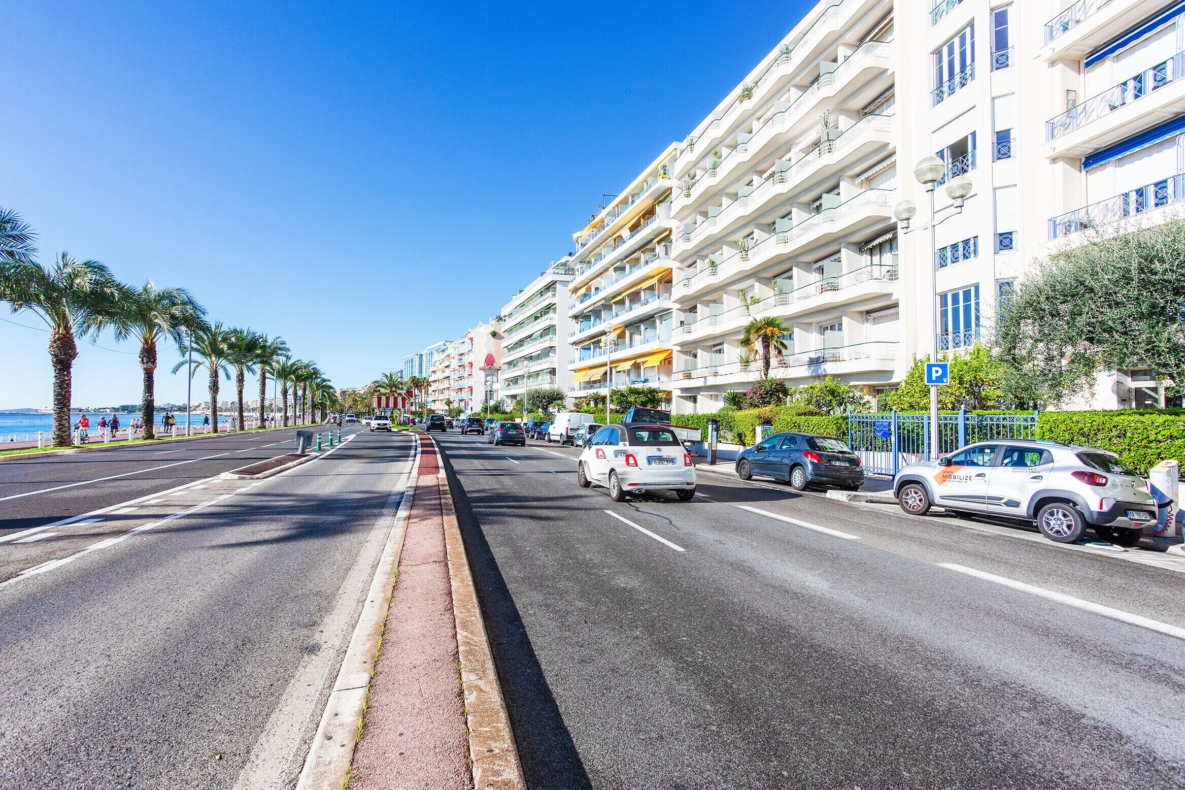 A coastal road lined with palm trees and multi-story buildings with balconies, a clear blue sky, and a parking sign.