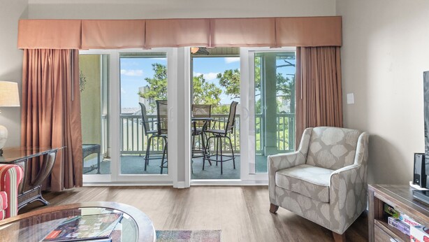 Ein Wohnzimmer mit Sofa, Couchtisch, TV-Schrank und Sessel. Eine Glastür führt zu einer Terrasse im Freien mit Tisch und Stühlen.