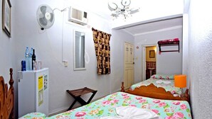 A bedroom with a bed, a small table, a chair, a fan, a refrigerator, and a door. - Hostal Casa Azul Santa Ana TRINIDAD (Trinidad)