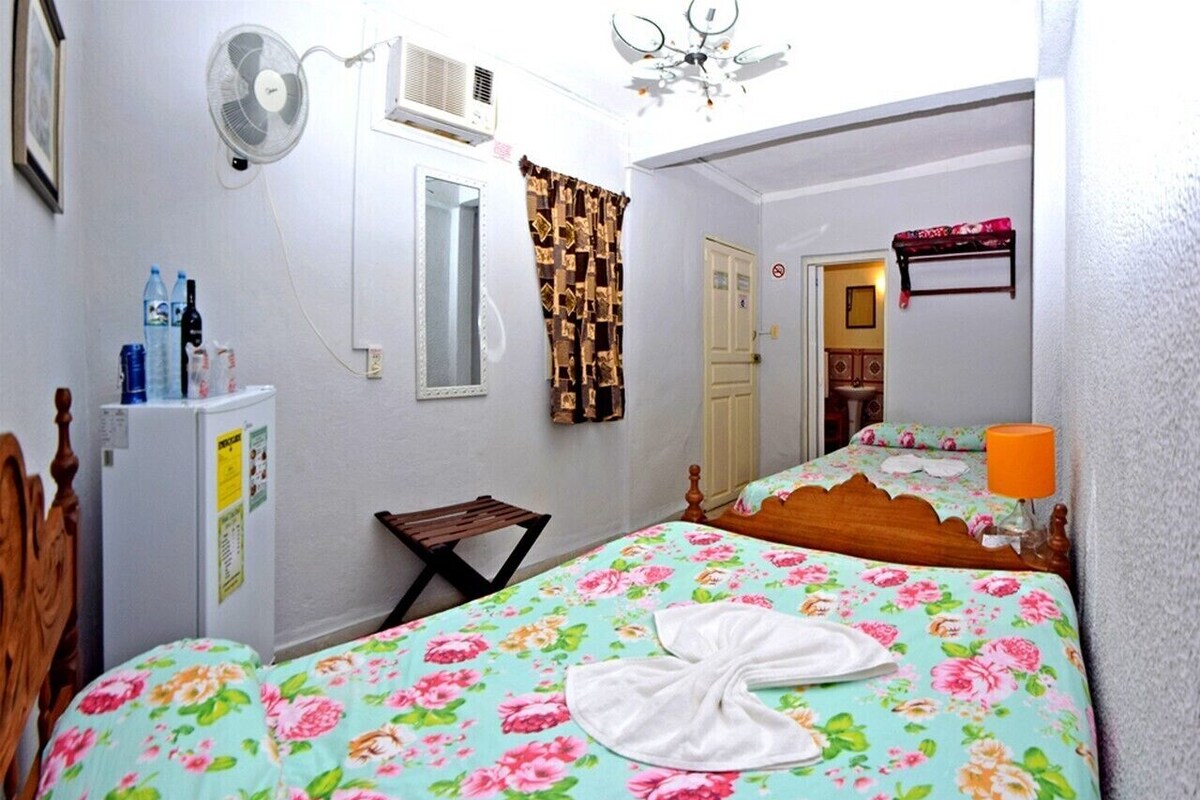 A bedroom with a bed, a small table, a chair, a fan, a refrigerator, and a door.