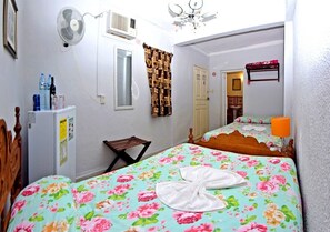 Un dormitorio con cama, una mesita, una silla, un ventilador, un refrigerador y una puerta.