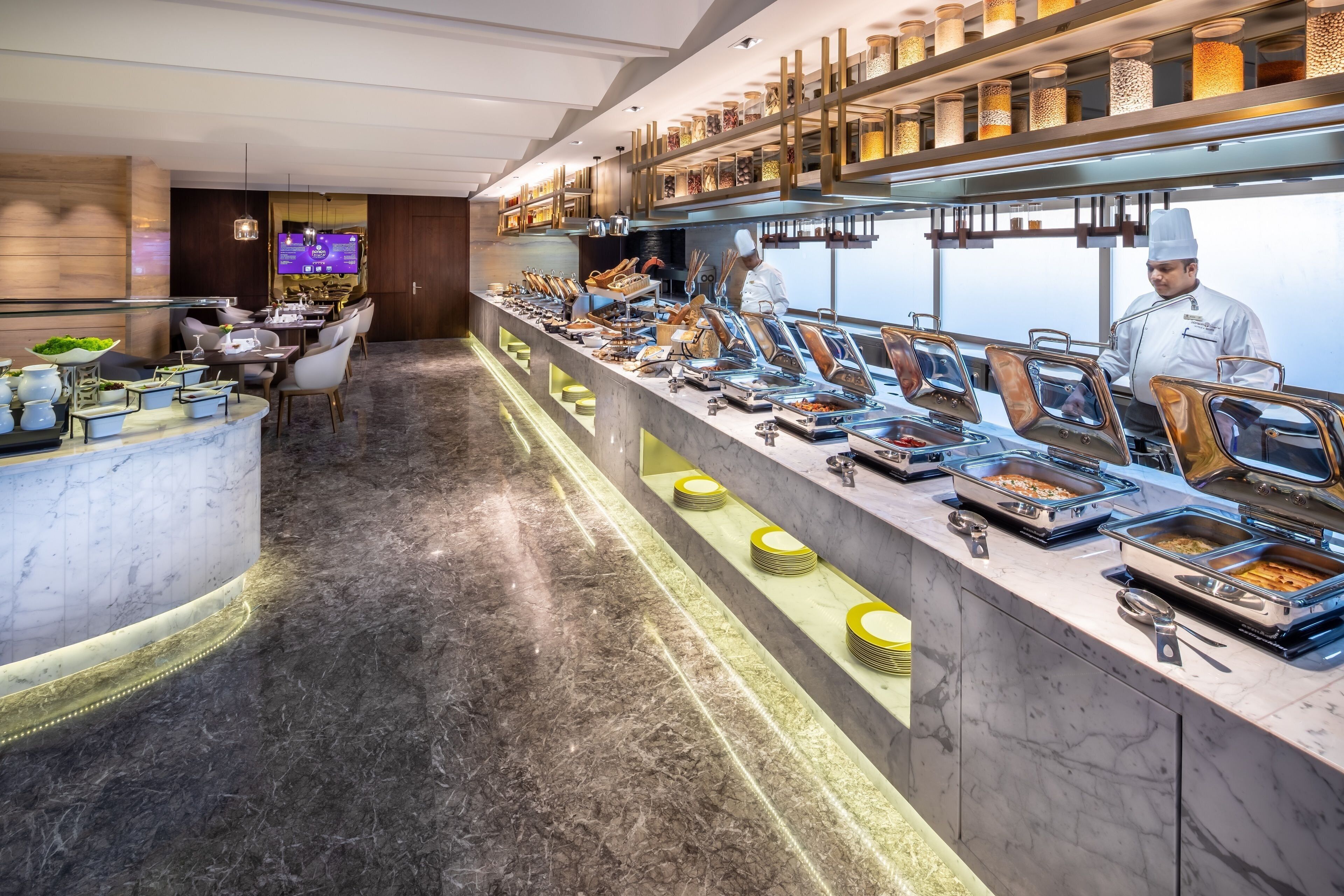 A buffet area with a marble floor, multiple serving stations, and a television screen in the background.