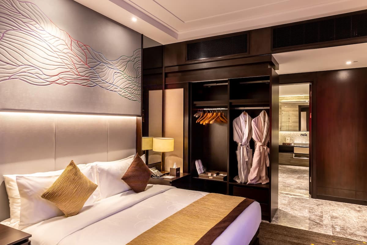 A hotel room with a large bed, a bedside table with a lamp, a wardrobe, and a bathroom visible through an open door.