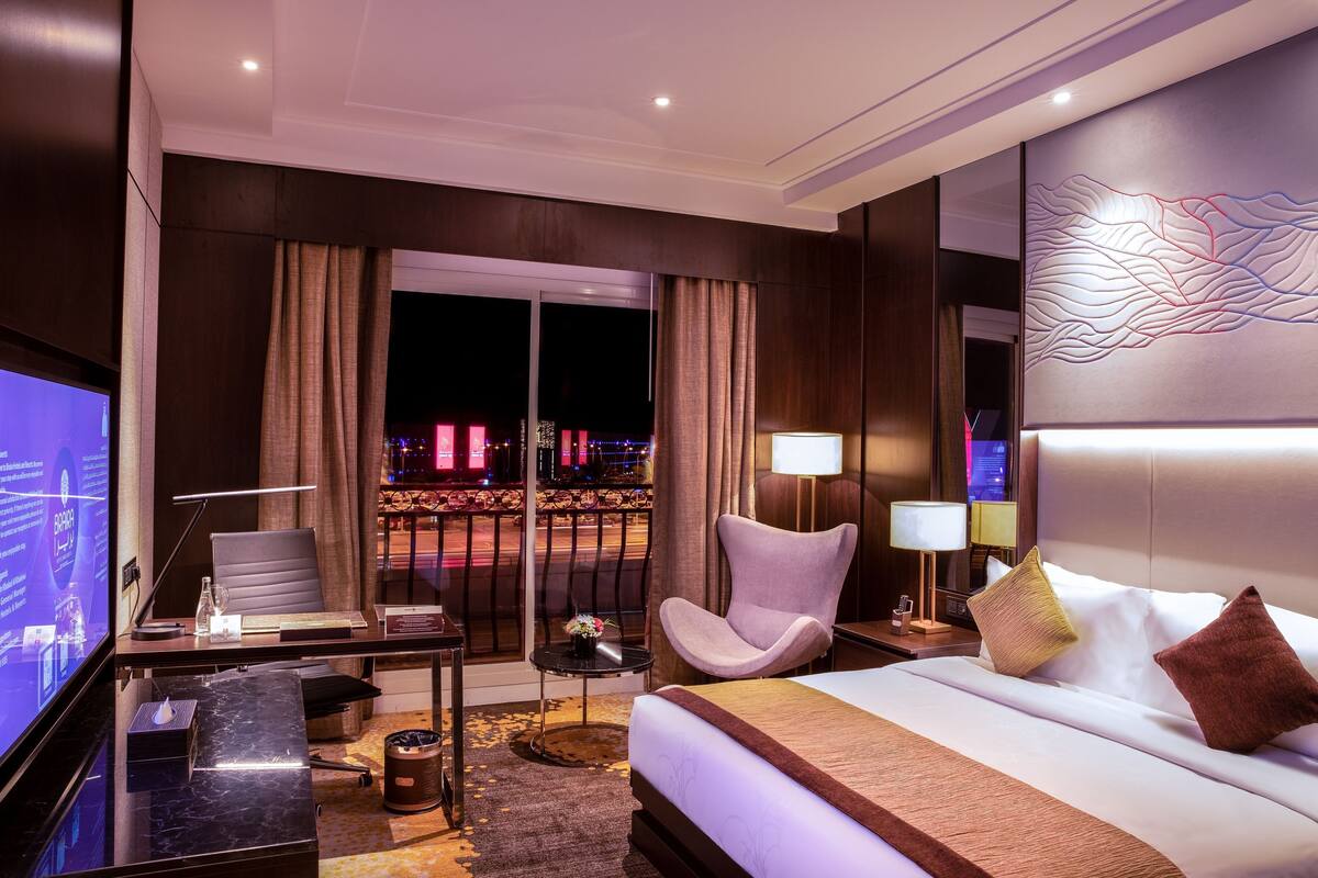 A hotel room with a bed, a desk with a computer, a chair, a TV, and a view of the city at night.