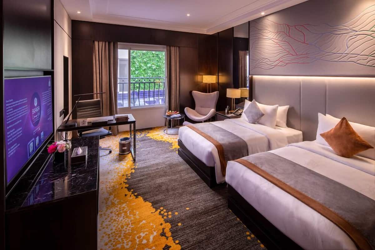 A hotel room with two beds, a desk, a TV, and a balcony with a view of greenery.