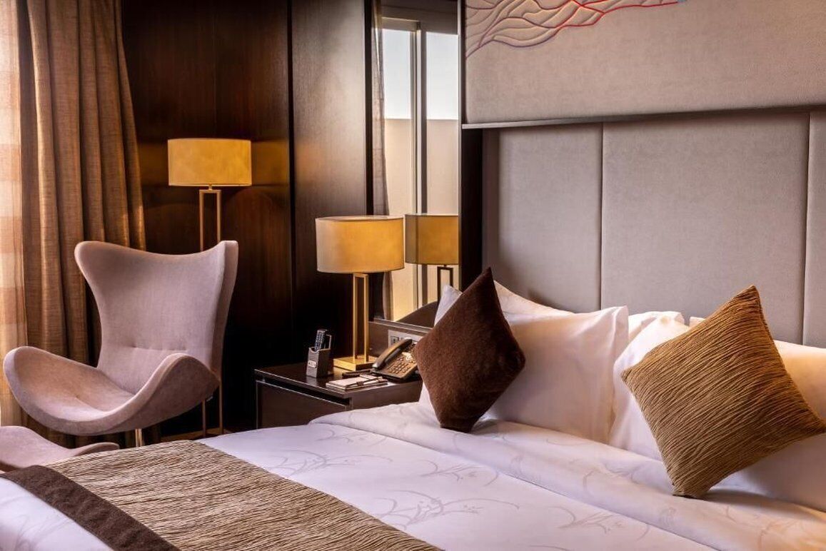 Una habitación de hotel con diseño moderno, que incluye una cama grande con ropa blanca, un sillón contemporáneo, dos lámparas de noche y un cabecero decorativo.