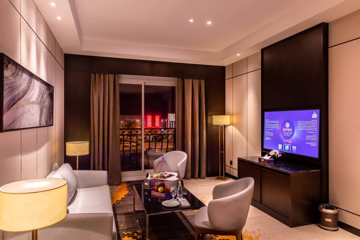 A modern hotel room with a large flat-screen TV, a sofa, a coffee table with fruit, and a view of a cityscape through the window.