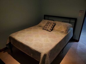 Una cama individual con un cojín estampado y otro liso, cabecero de metal y suelo de baldosas.