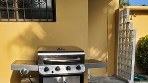 Una parrilla de acero inoxidable con cuatro quemadores, una repisa lateral y un armario de almacenamiento, ubicada en un patio de piedra.