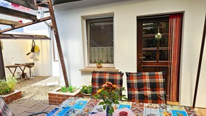 Une terrasse avec une table recouverte d’une mosaïque de carreaux aux motifs marins, deux chaises à carreaux, un vase rempli de fleurs et une échelle en bois appuyée contre un mur blanc.