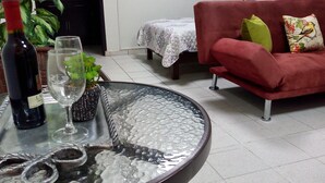 A living room with a round glass coffee table, a red sofa, a small table with a bottle of wine and glasses, and a potted plant.