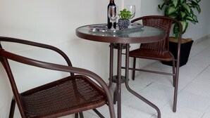 A dining area with two wicker chairs and a round table, a bottle of wine, and two glasses. There is a potted plant to the right and a sign on the wall.